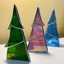 Three triangular stained glass Christmas tree ornaments in green, pink, and blue, arranged on a white surface against a pale yellow background.