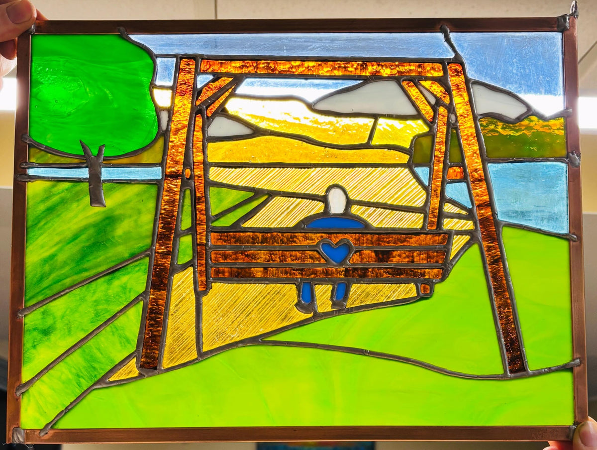 A stained glass art piece depicting a person sitting on a swing in a park, held up by hands. The design features a bright green grassy field, a yellow path, a blue sky, and a brown swing frame with a figure in blue.