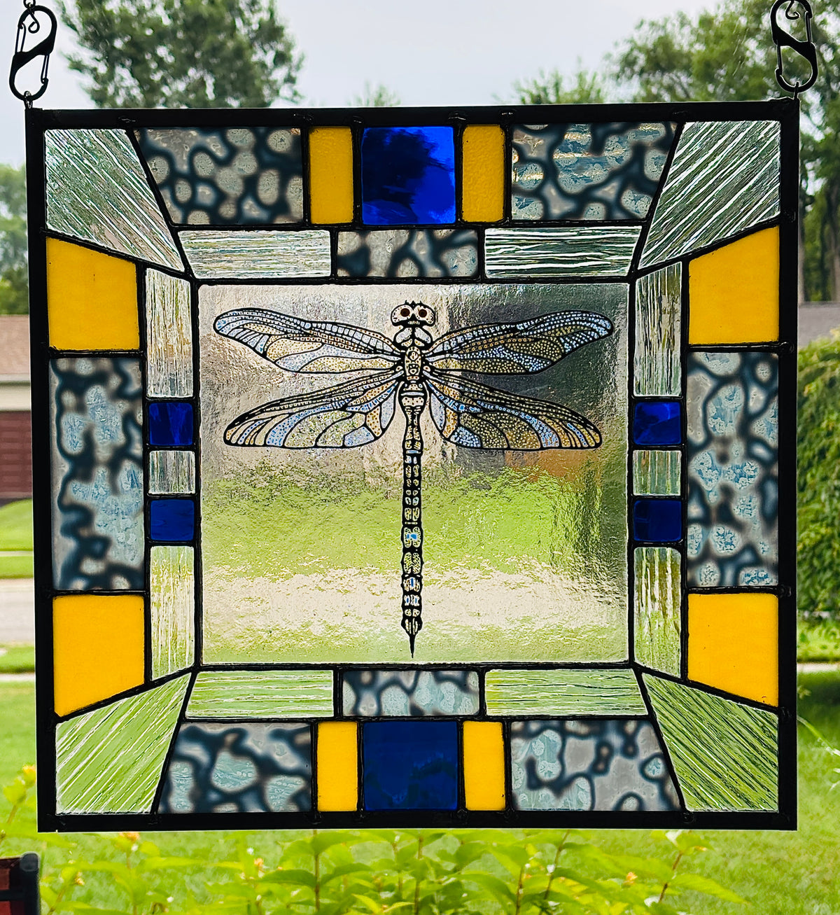 A stained glass window panel featuring a detailed dragonfly design in the center, surrounded by geometric patterns in blue, yellow, green, and textured gray, framed by black metal bars and hanging from black metal hooks.