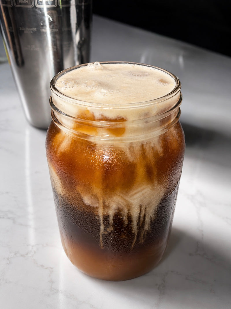 An iced coffee drink made from Verity's Cold Foam Powder, in a clear glass mason jar with condensation, layered with dark coffee and creamy milk, topped with foam, set on a light marble countertop with a metal shaker in the background.