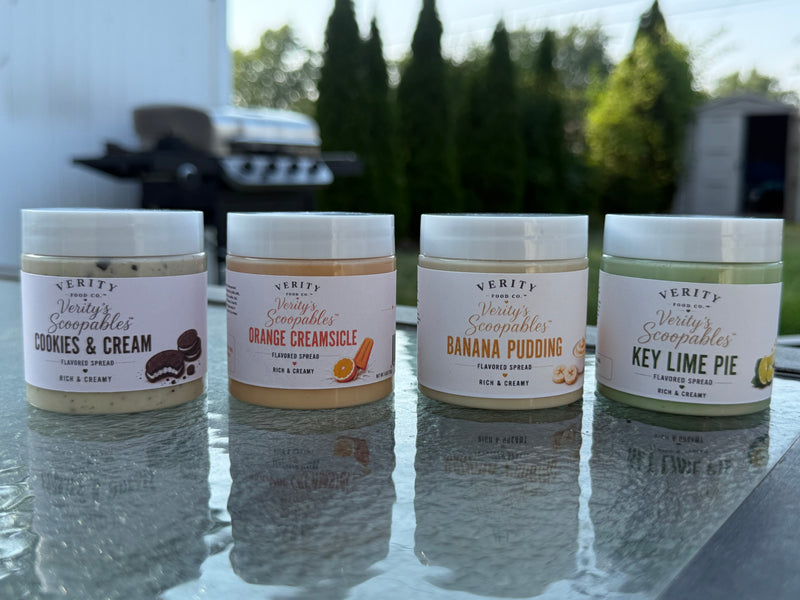Four jars of Verity Food Company 'Verity's Scoopables' flavored spread arranged in a row on a glass patio table outdoors, with blurred trees and a grill in the background. The jars are labeled Cookies & Cream, Orange Creamsicle, Banana Pudding, and Key Lime Pie.