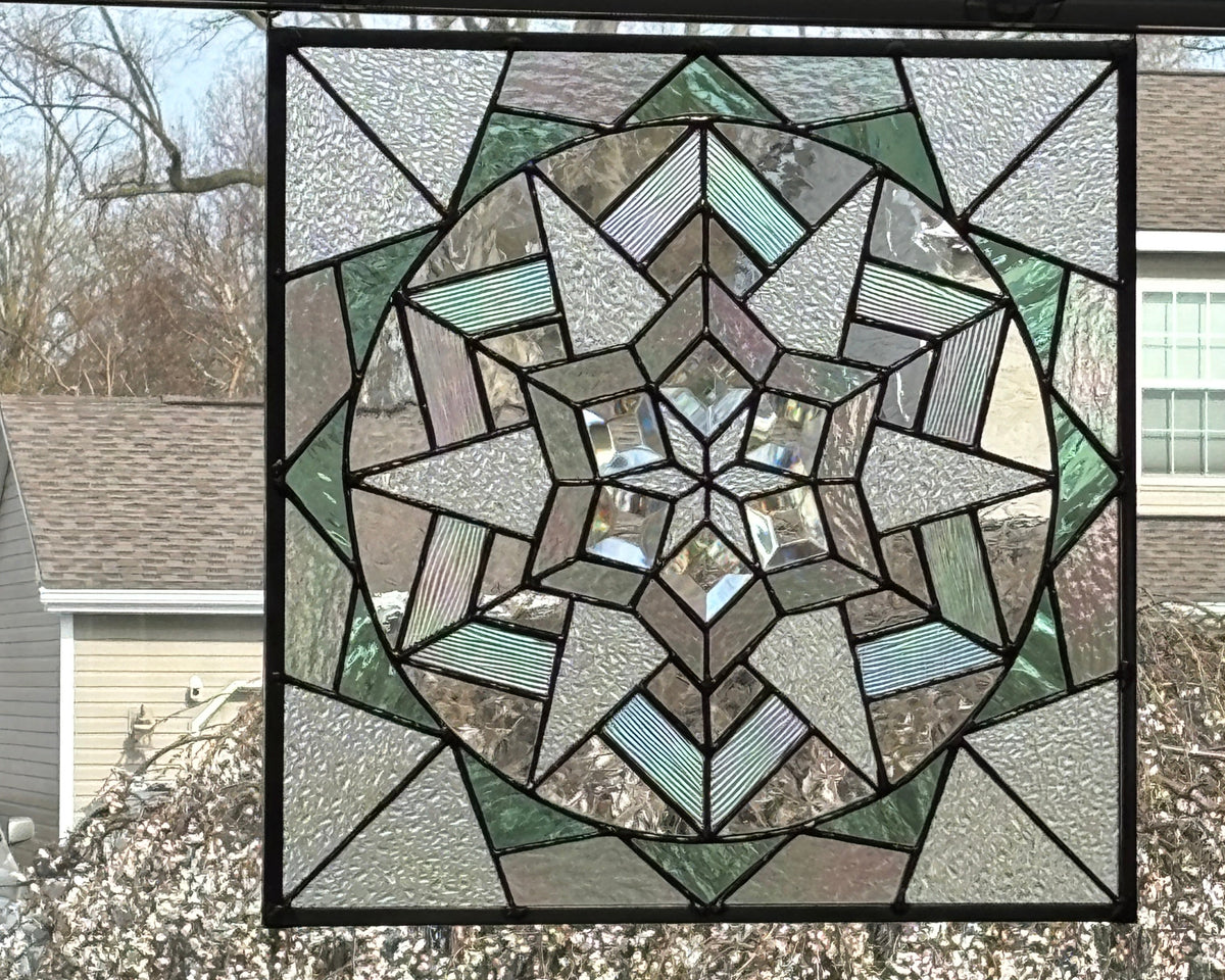 A square stained glass window panel with a geometric starburst design, featuring textured gray, green, and clear glass sections outlined in black metal, shown hanging on a window with a residential outdoor view visible.